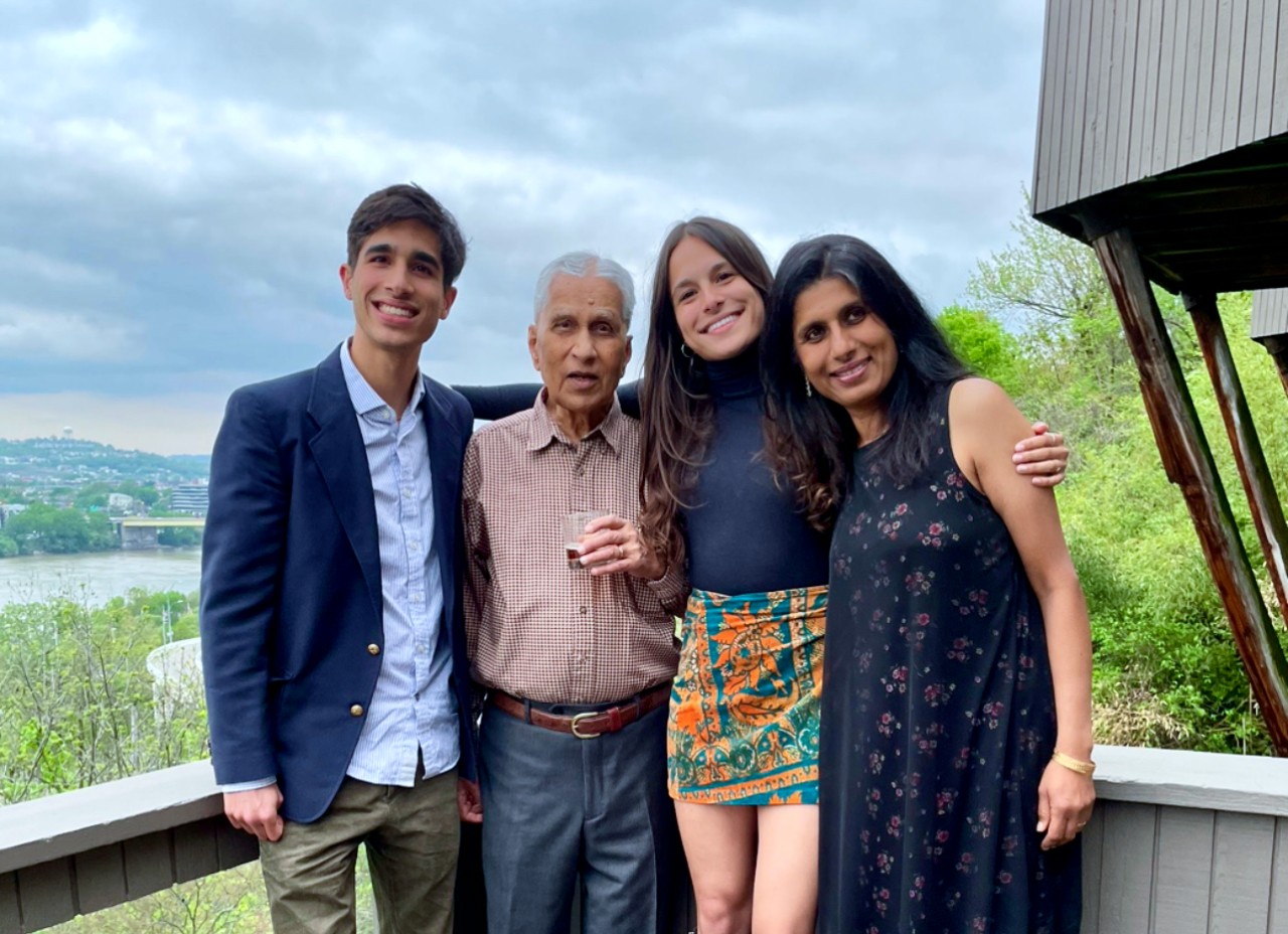 Two men and two women stand together facing forward on a balcony with a river in the background.