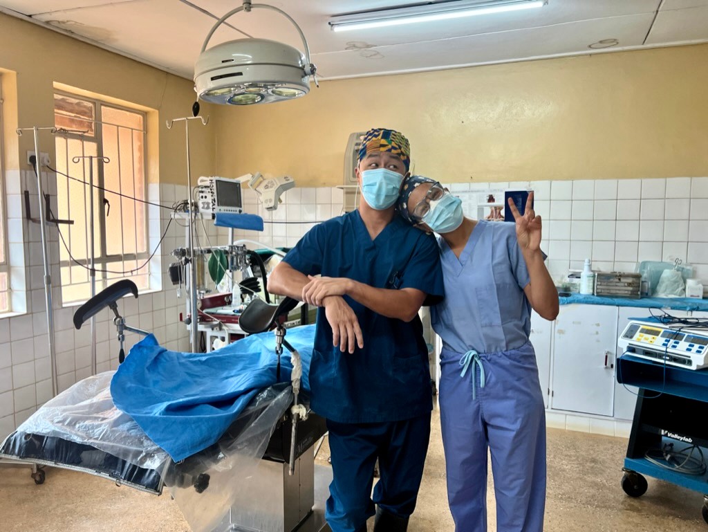 Two doctors stand together in an operating room wearing surgical masks and scrubs.