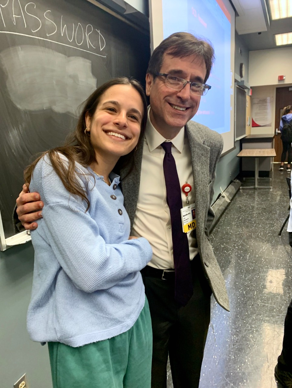 Young woman on left stands with a man in front of a blackboard in a college classroom.