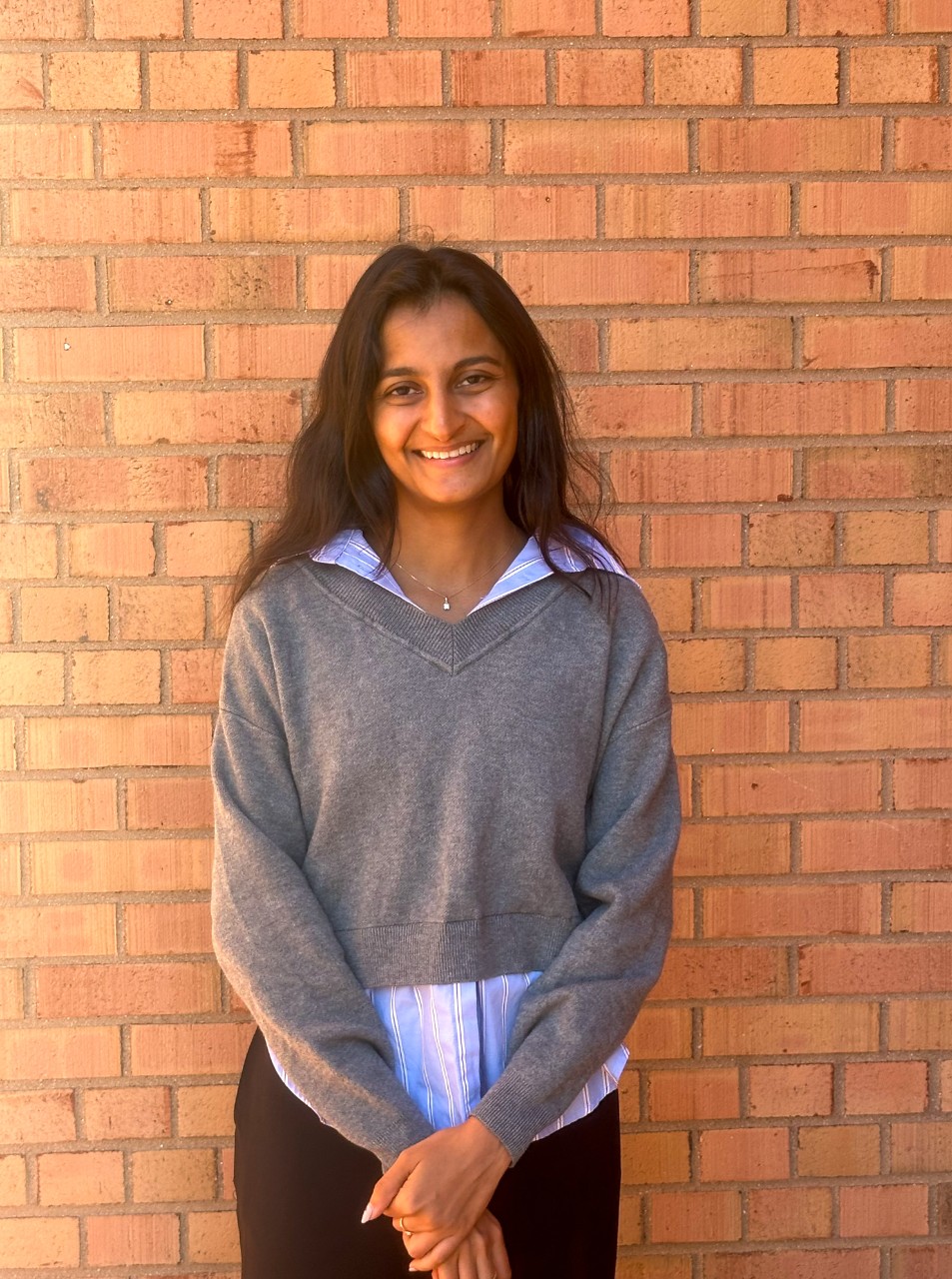 A woman in a gray shirt stands in front of a brick wall and smiles. 