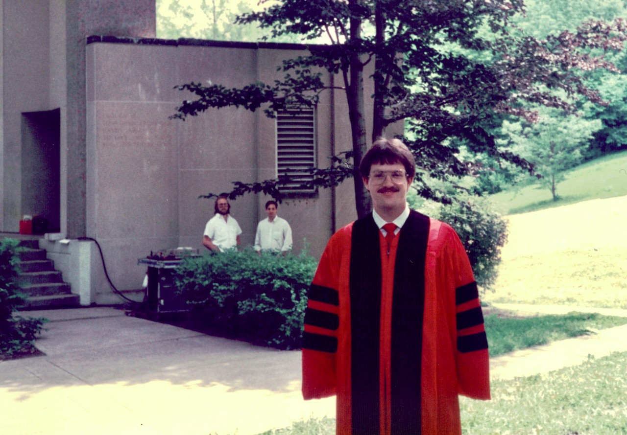 Man wearing graduate degree cap and gown stands in front of UC's College of Medicine with two men standing int the background.
