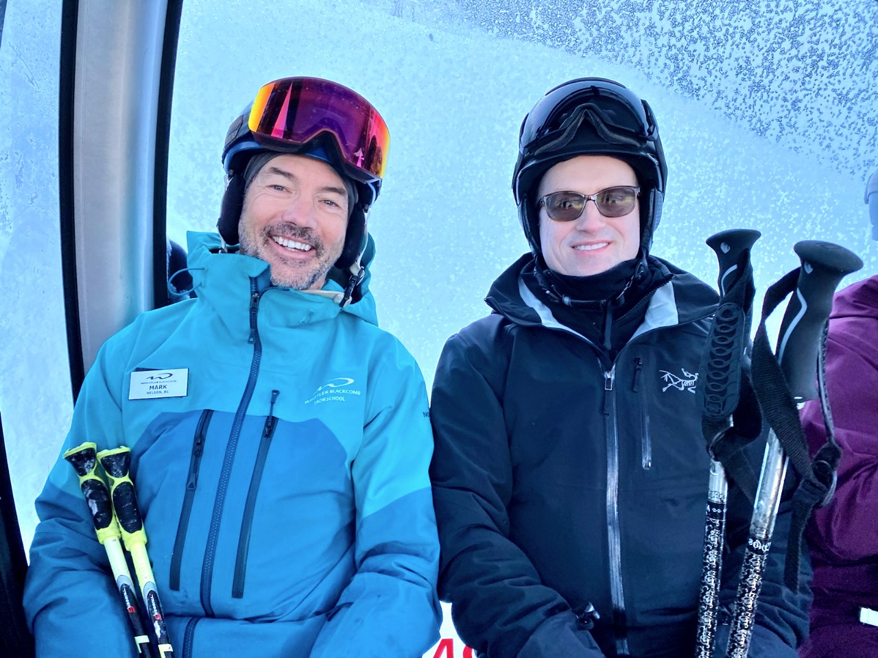 Two men face forward while sitting in a ski lift wearing goggles and holding ski poles.