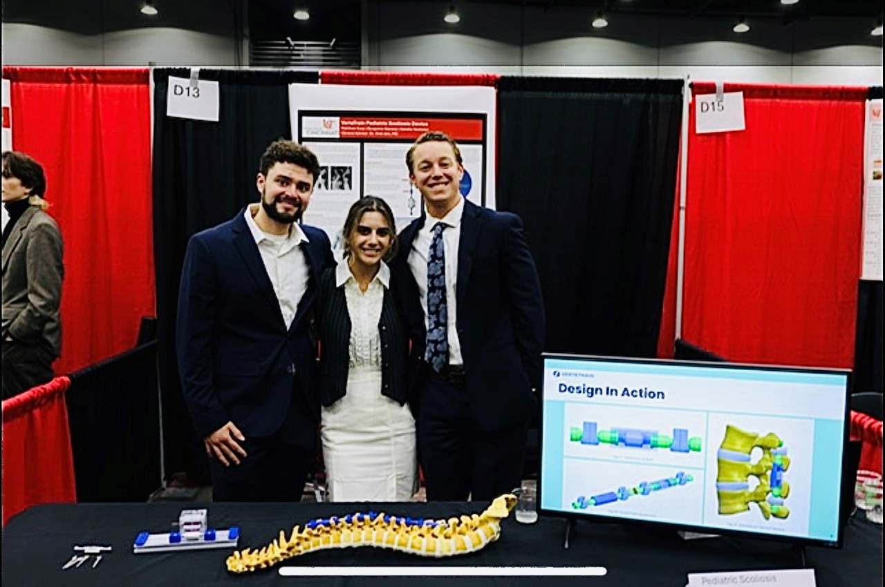 Three people pose for a photo in a booth at a conference with a computer screen in front of them.