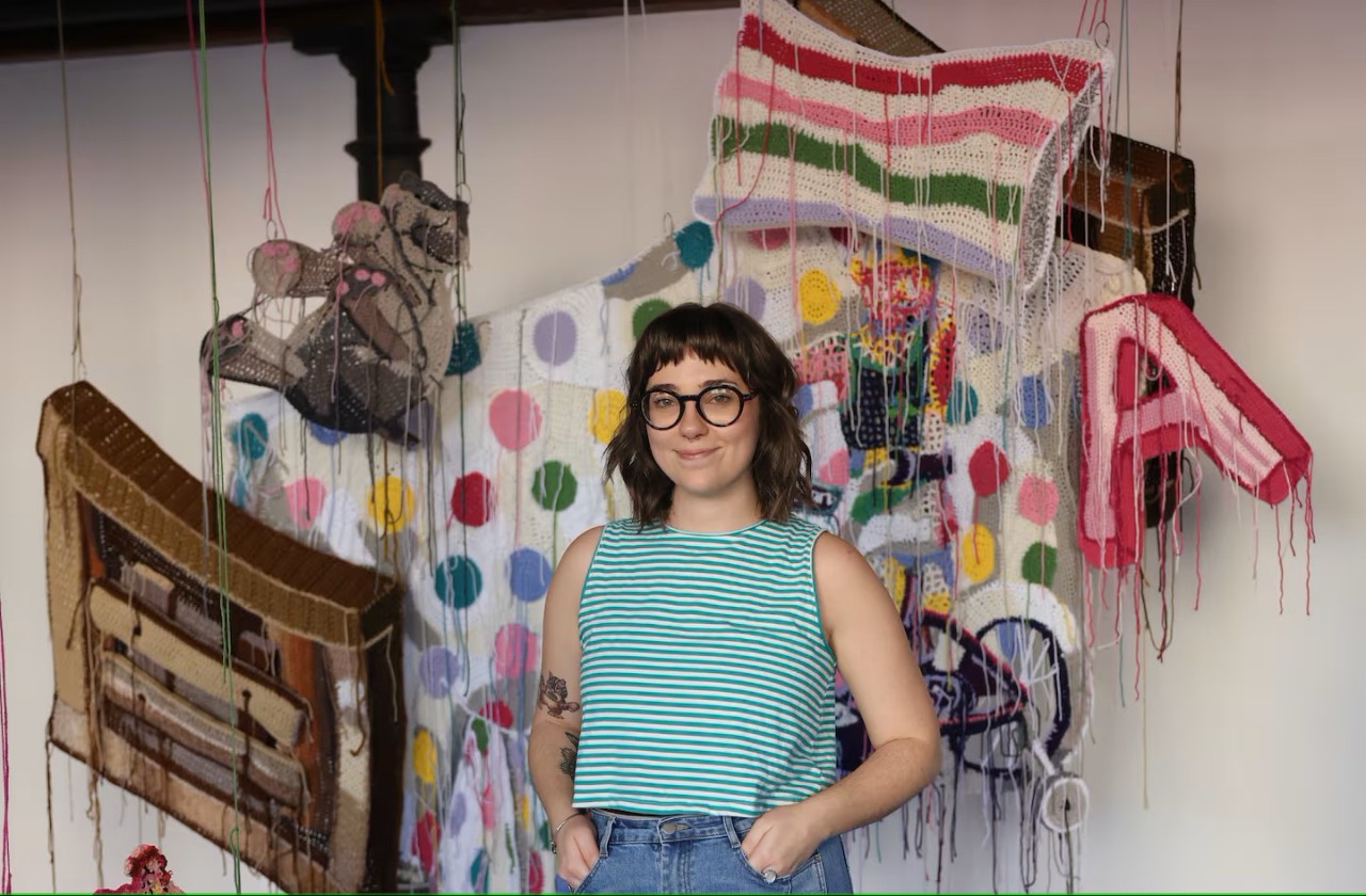 Artist Baylee Schmitt standing in front of her yarn artworks which are scale representations of her childhood home 