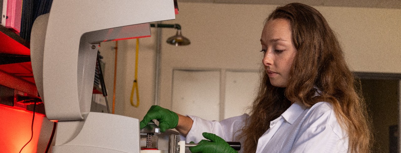 Katherine Schlaak working in the lab with testing equipment. She wears a lab coat and green rubber gloves. 
