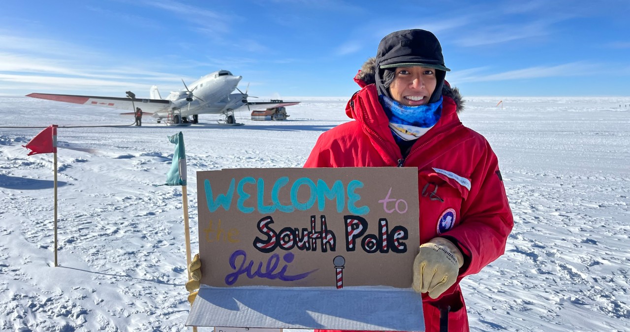 Julianne Fernandez wearing a NOAA jacket, gloves and hat holds up a sign reading 'welcome to the South Pole, Juli!' on the ice in front of a plane on snow skids in Antarctica.