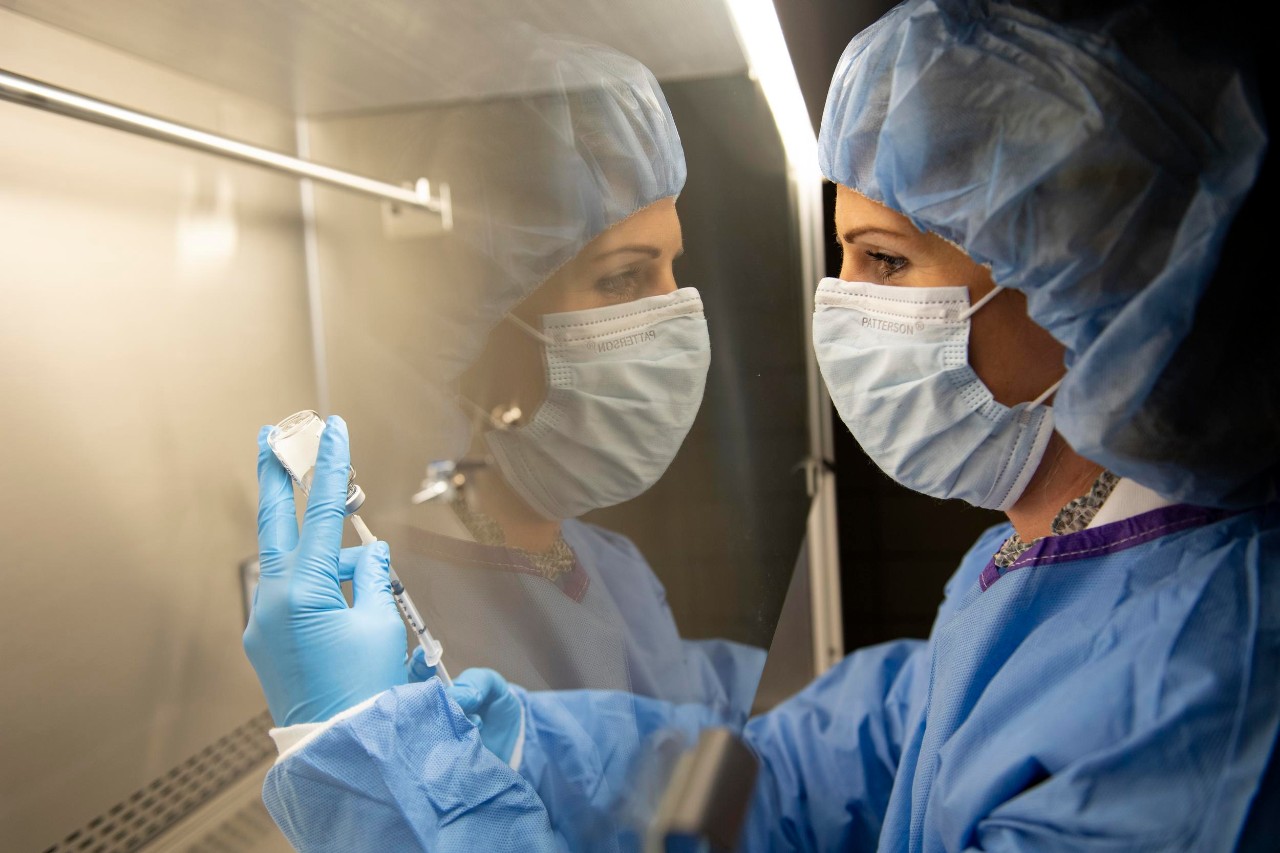 A UC College of Pharmacy student in a facemask and gloves draws a syringe under a glass hood that reflects her image.