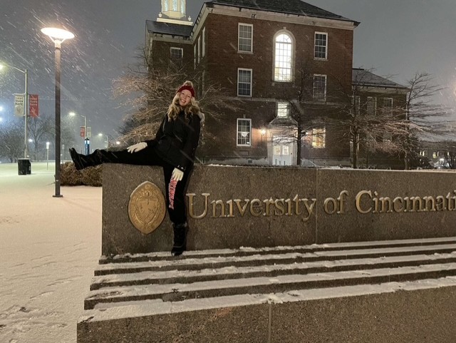 dressed in winter clothes Elisse Martin sits on a University of Cincinnati sign as snow falls