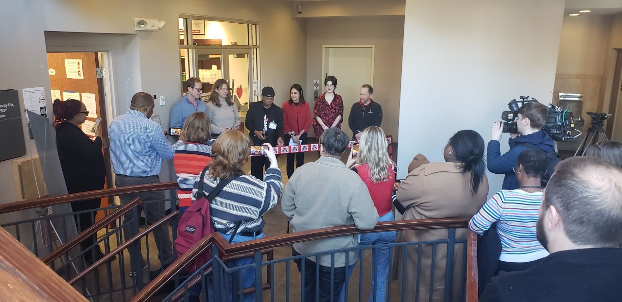 several people gather in front of pantry center as 6 individual hold ribbon as Daniel cummins cuts it