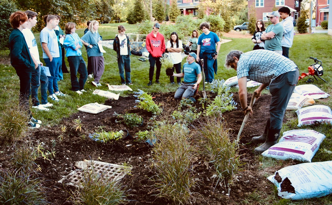 Several students and faculty wearing boots and holding shovels dig into soil while planting a rain garden.