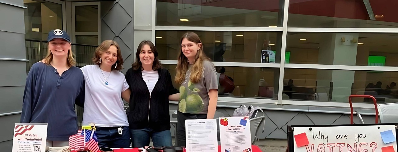 Four UC Votes student workers registering students to vote stand next to a whiteboard with the question "Why are you voting?" and pose for a photo in front of their table outside of TUC.