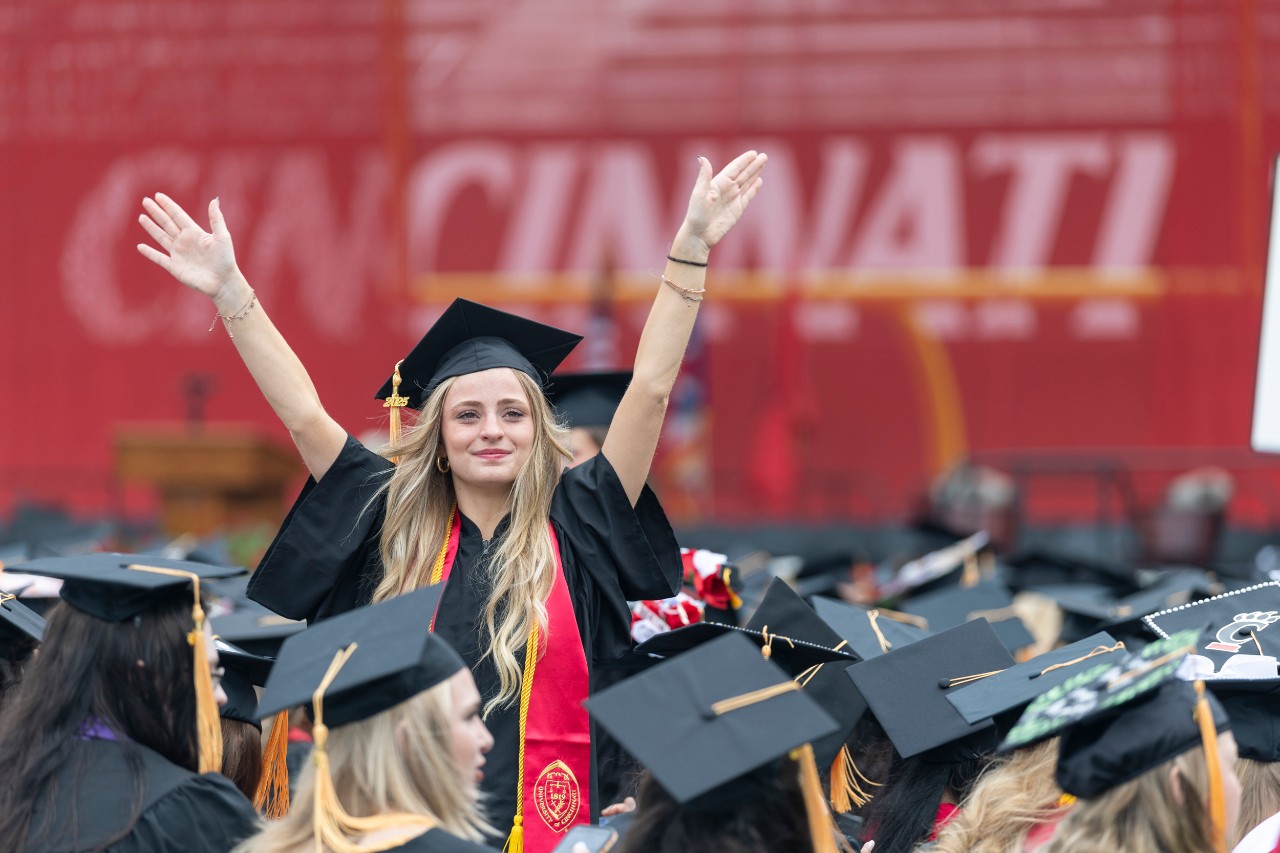 A UC grad holds up her arms in a sea of black caps and gowns at Nippert Stadium.