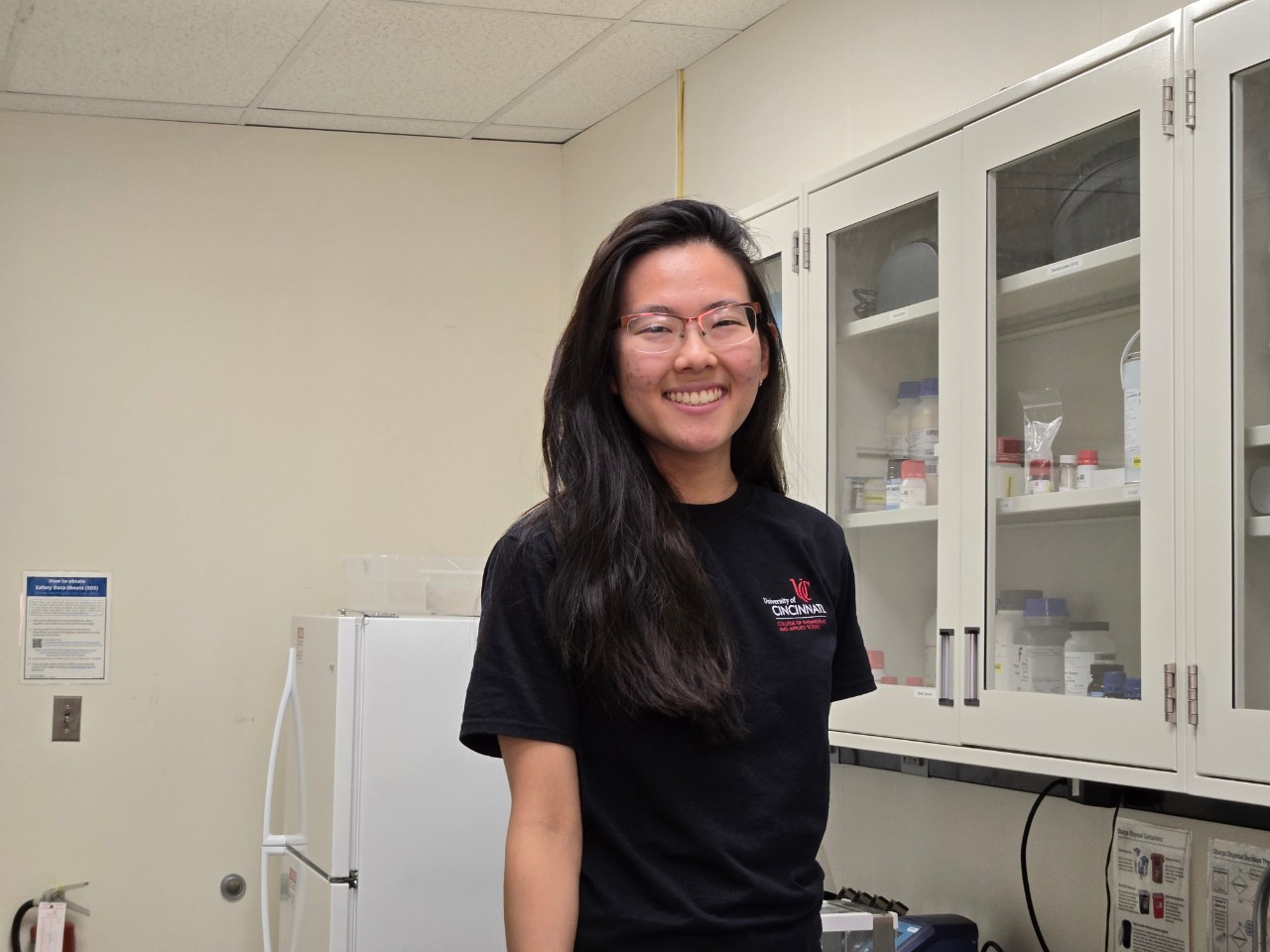Karen Noda Morishita stands in a University of Cincinnati research lab and smiles at at the camera. She wears a black shirt with the UC logo on it. 