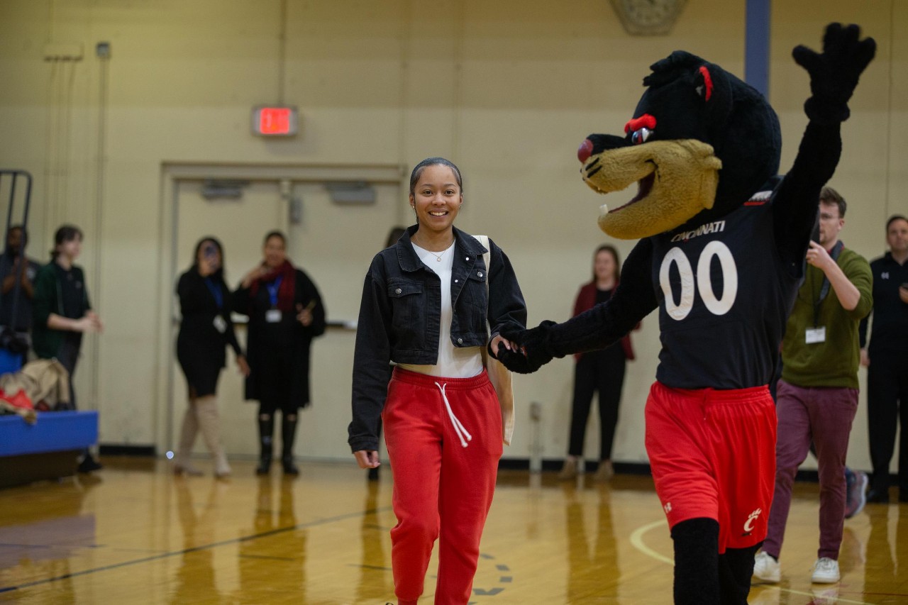 A high school student, recently accepted into college, is lead across a basketball court with the UC Bearcats mascot next to her.