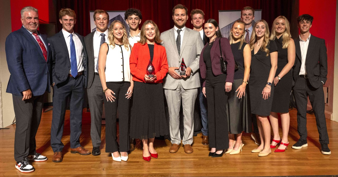 Distinguished Service winner Shenan Murphy stands at the far left of a group photo with students after the real estate dinner. All in professional dress, standings on a wooden stage.