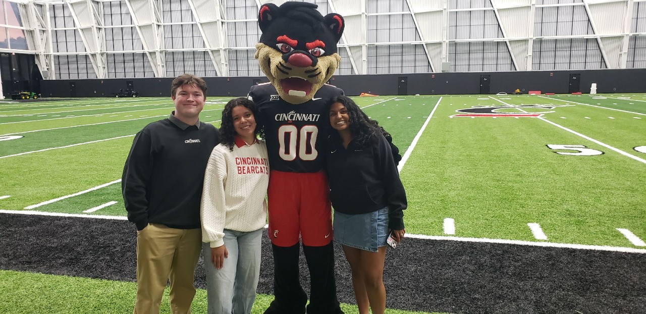 Josh Copley, Wilaini Alicea, Kripa Kumaran and the Bearcat mascot shown on indoor football field in Sheakley sports complex