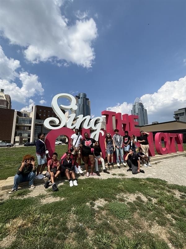 Several Cincinnati Ambassador students shown sitting near a Cincy Sing sign in downtown Cincinnati