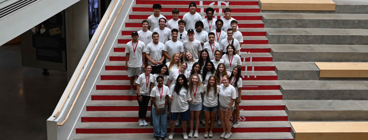 The 2025 Lindner Business Immersion Camp cohort poses in white t-shirts on the atrium stairs inside Lindner Hall.