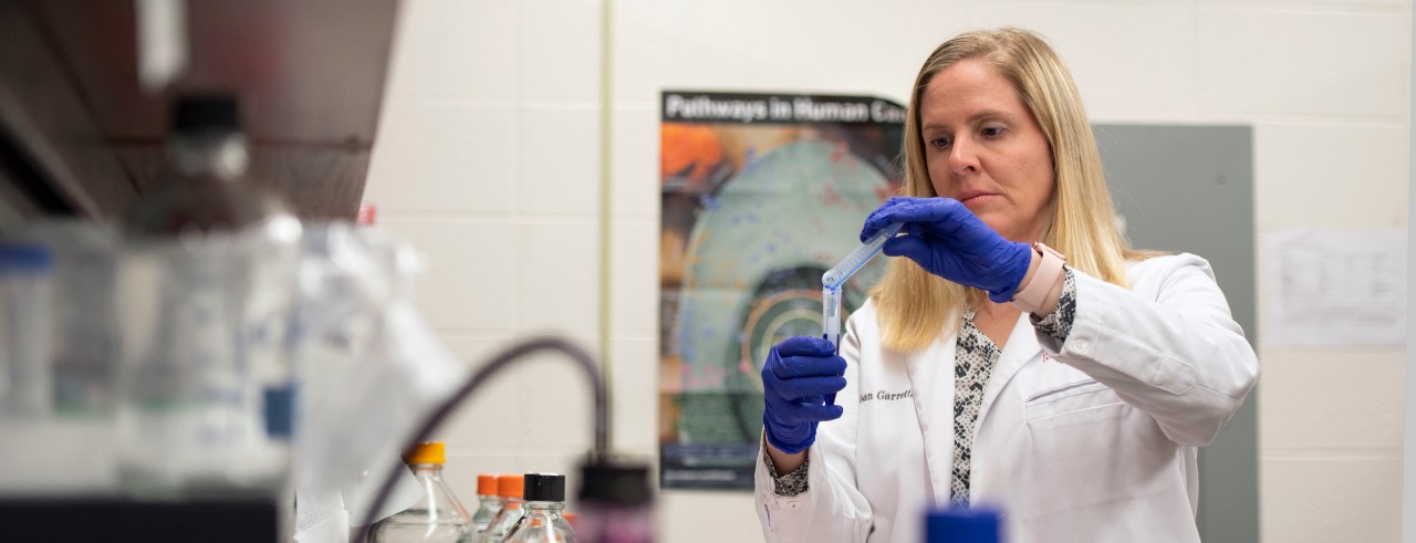 Joan Garrett pipettes a sample in a test tube while wearing a white lab coat and blue rubber gloves in the laboratory