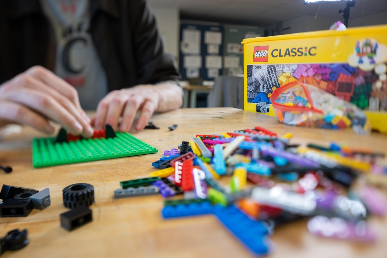 See the hands and torso a student arranging a pile of lego bricks on a desk
