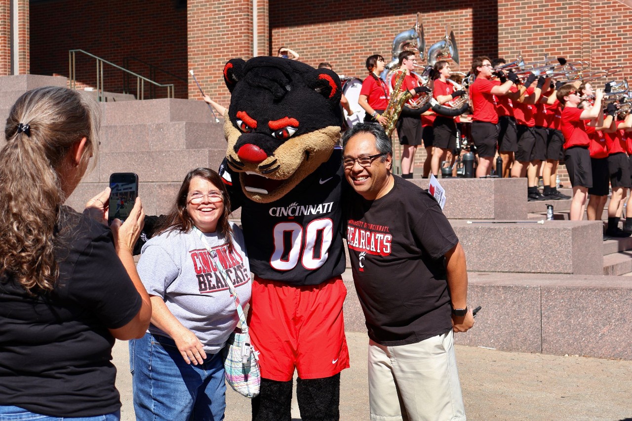 Mom and dad stand next to the Bearcat Mascot while a daughter takes a photo and the Bearcat Band is seen playing in the background