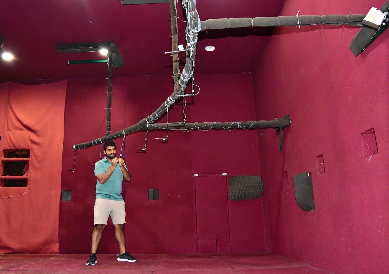 Kaurab Gautam stand sin an empty room in front of a microphone array. The walls, floor and ceiling are covered in foam padding.