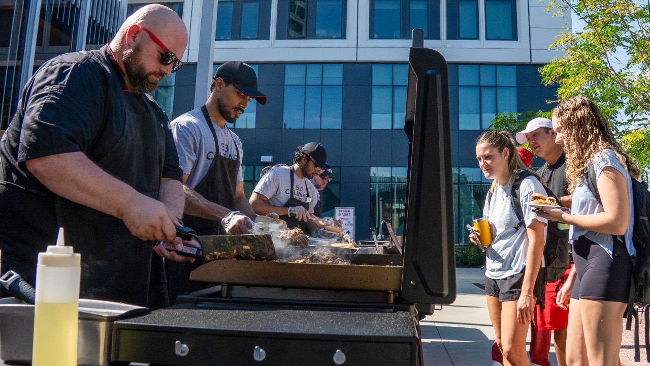 Four men shown at big grills tossing meat and three hungry students watch nearby waiting for a plate