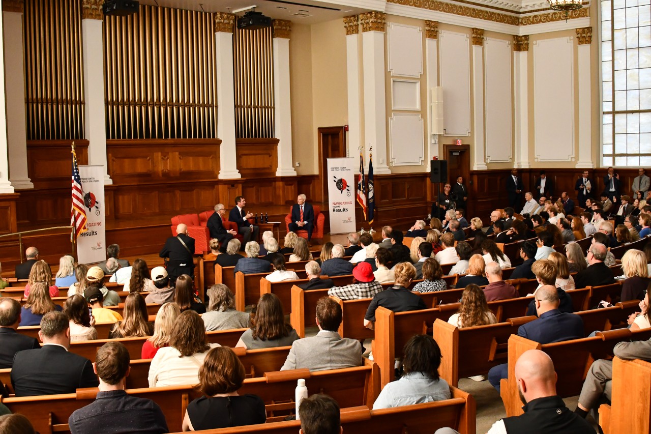 A large crowd of spectators listens to speakers seated on stage at Probasco Auditorium.