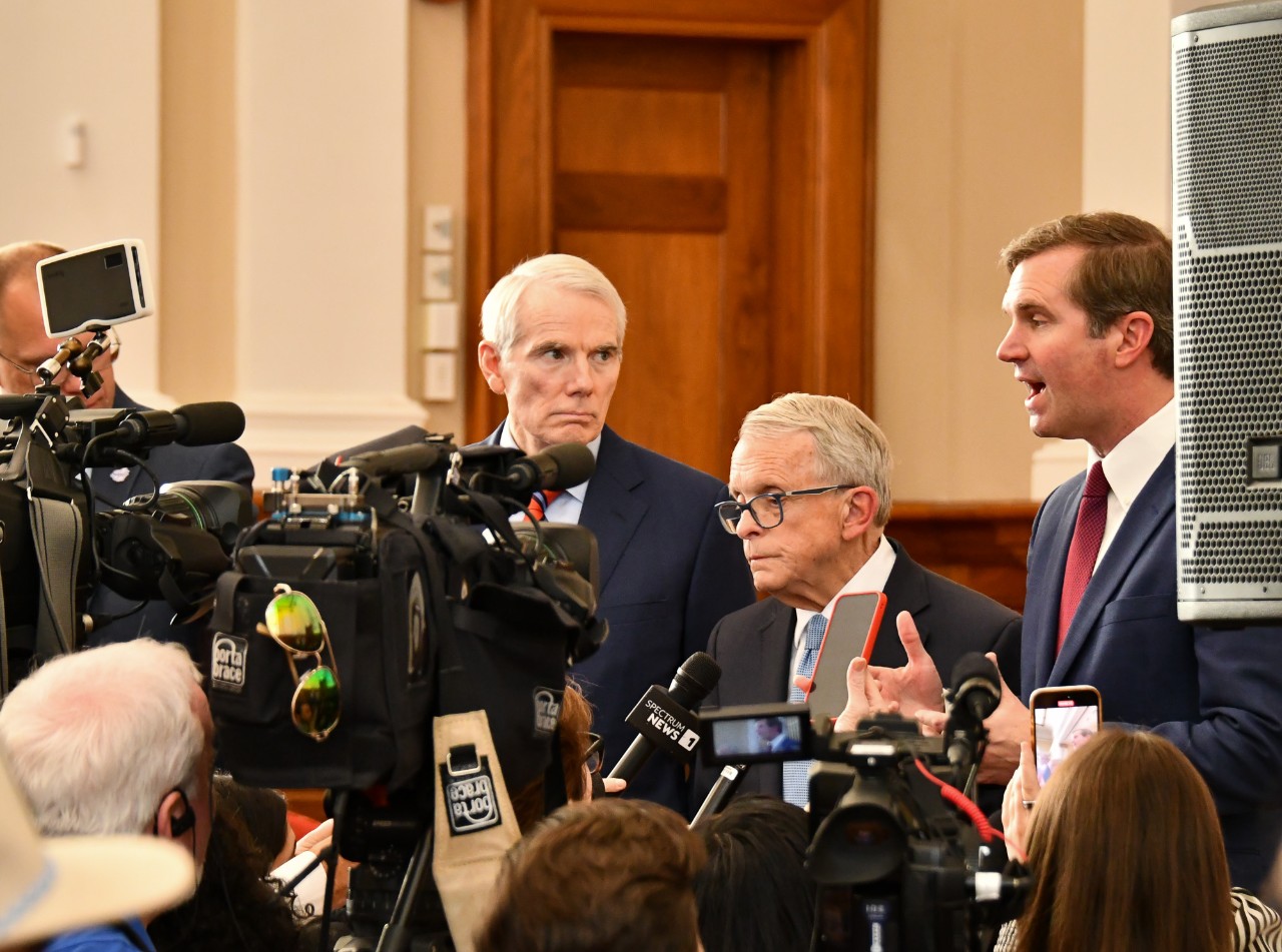 Rob Portman, Mike DeWine and Andy Beshear stand in front of a scrum of news cameras.